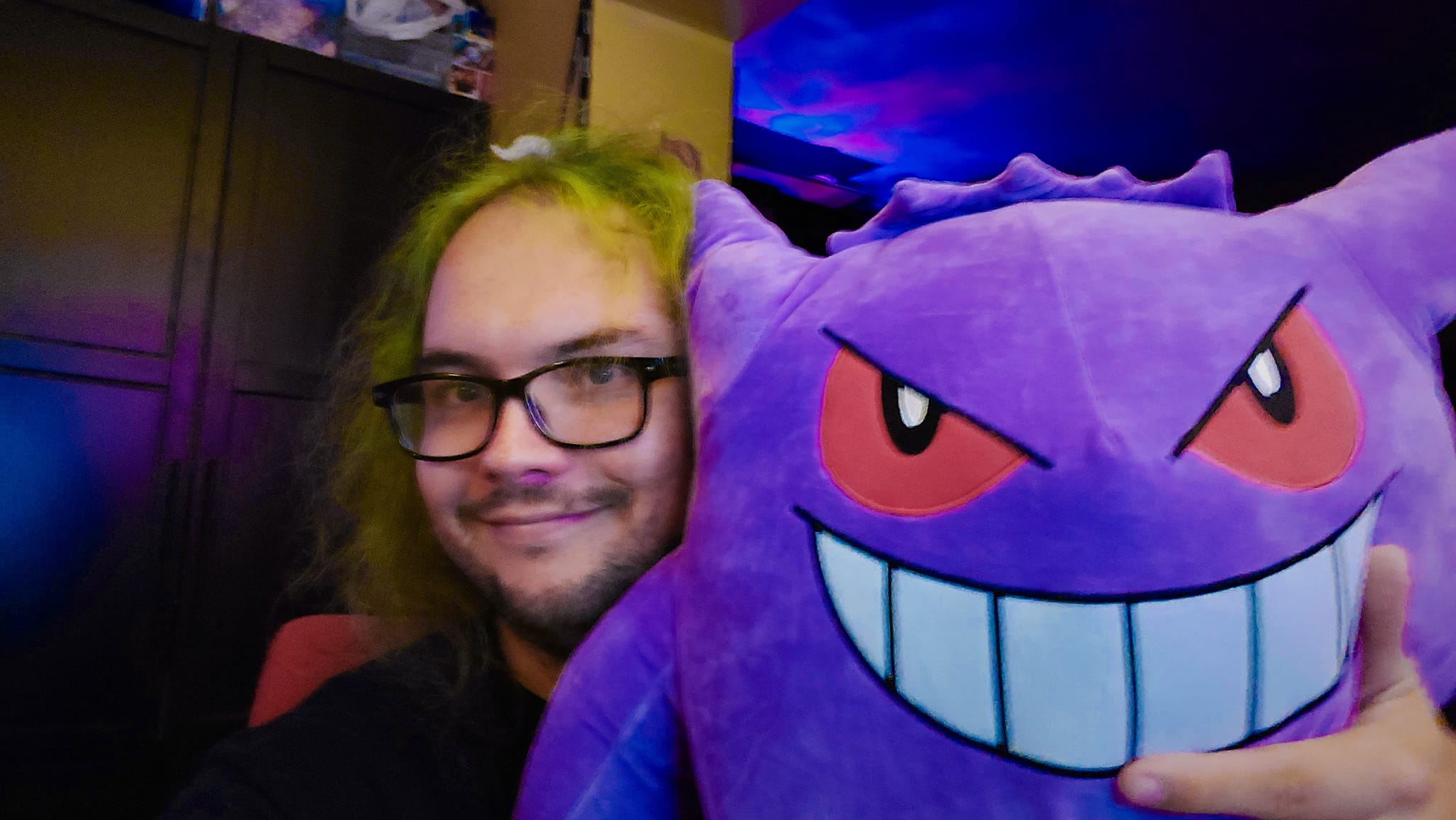 A man with green hair sits in a computer chair inside, with larged stuffed Gengar on his lap